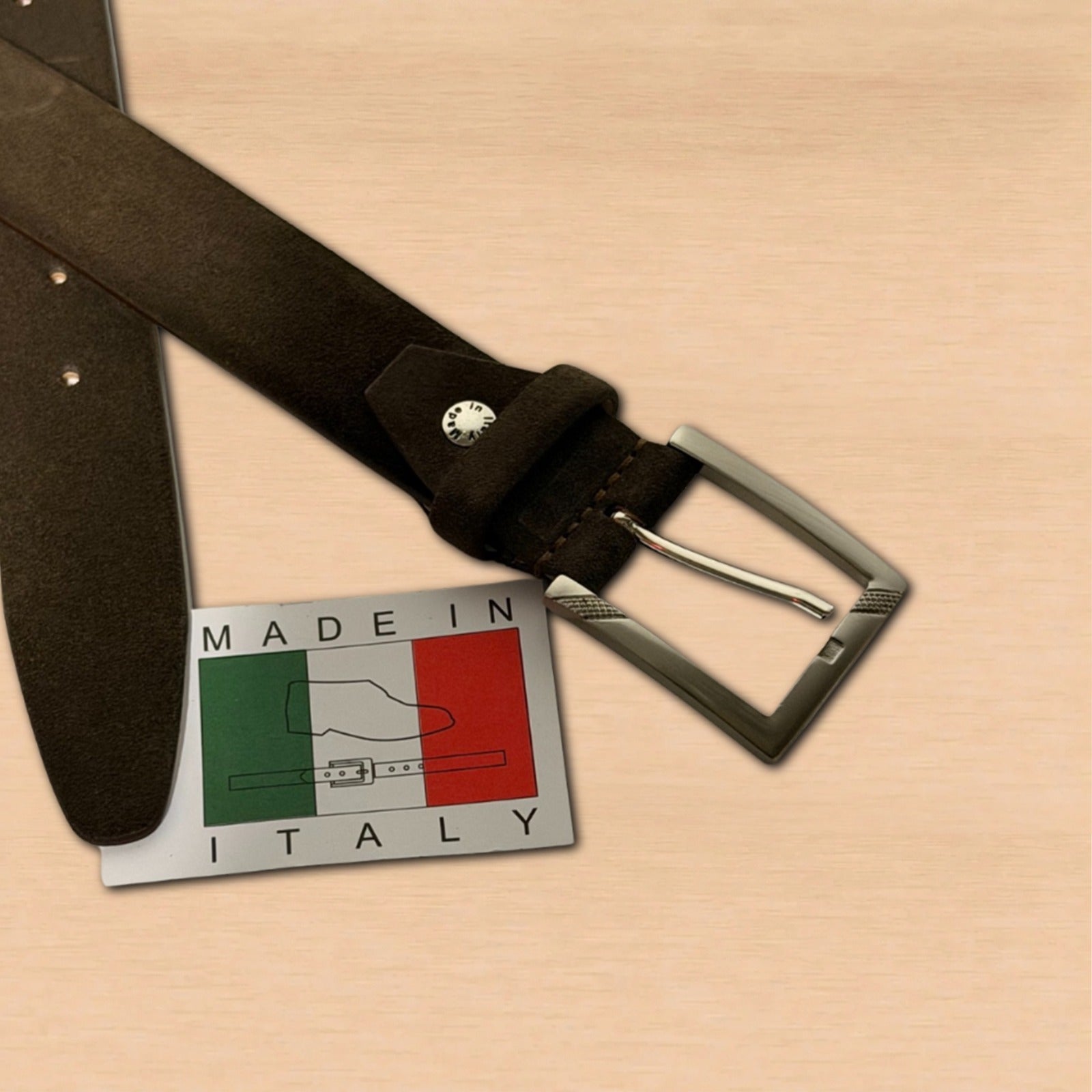 A dark brown suede belt with a silver metal buckle, displayed on a light wood background next to a silver 'MADE IN ITALY' tag featuring the Italian flag.