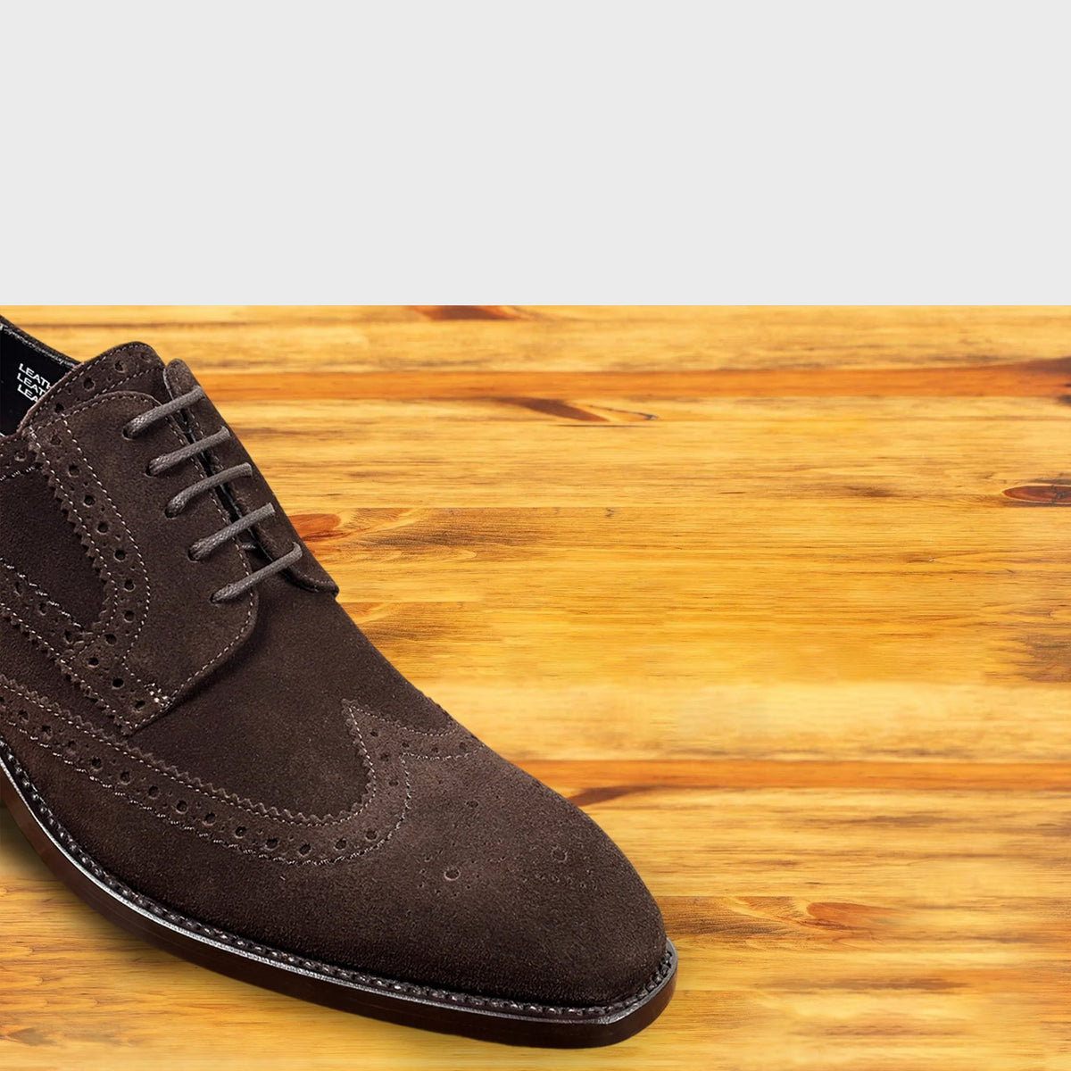 A  brown suede wingtip oxford shoe with brogue detailing and matching laces, positioned on a light-colored wooden surface.