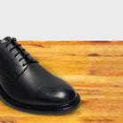 A close-up of a black leather lace-up dress shoe positioned on a wooden surface.