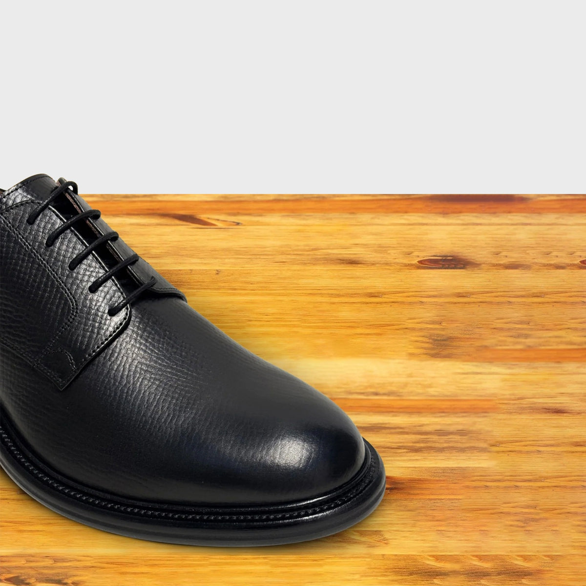 A close-up of a black leather lace-up dress shoe positioned on a wooden surface.