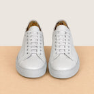 A pair of light white leather low-top sneakers with matching laces, shown from the front on a light wood surface against a cream background.