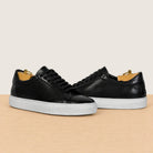 A pair of black leather low-top sneakers with textured perforated uppers, black laces, and thick white platform soles, displayed on a light wood surface against a warm beige background.