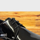 A close-up of a glossy black dress shoe with black laces, photographed on a warm wood surface.