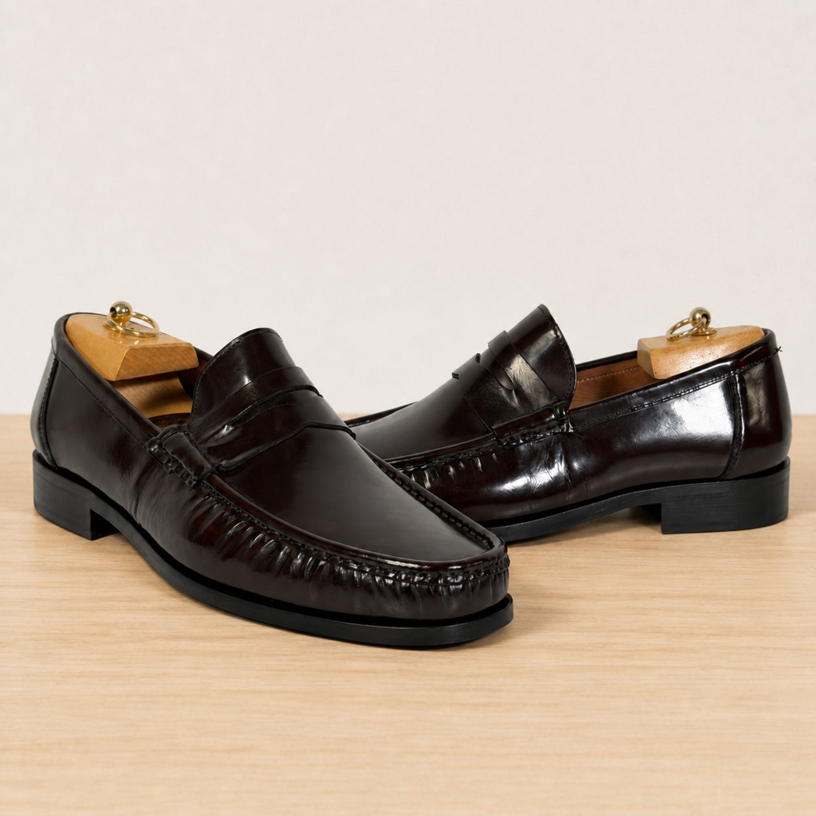 A pair of burgundy glossy leather penny loafers with textured moccasin-style stitching around the toes, displayed on wooden shoe trees on a light wood surface against a neutral beige background.