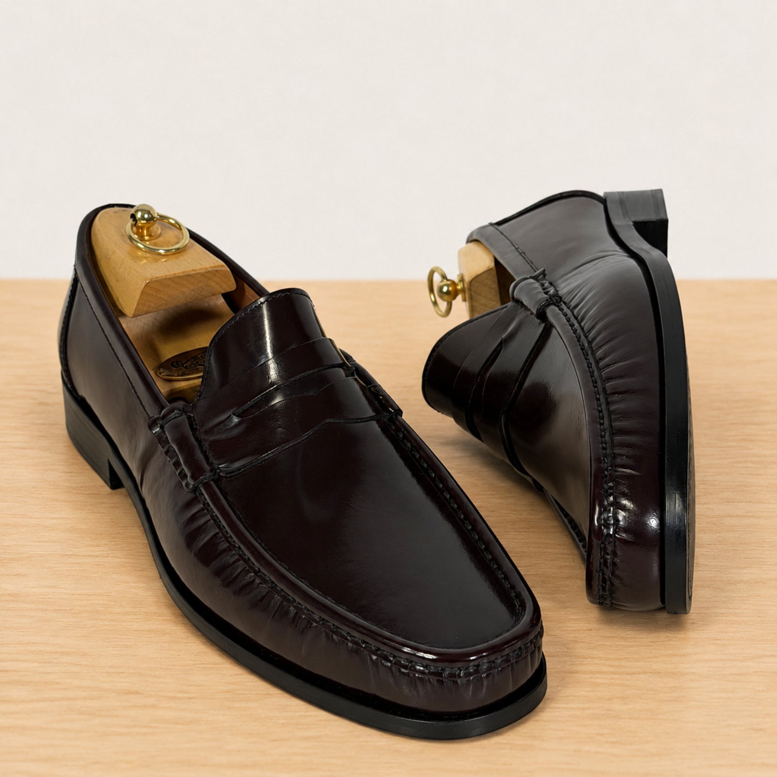 A pair of burgundy glossy leather penny loafers with wooden shoe trees inside, displayed on a light wood surface against a neutral off-white background.