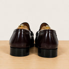 A pair of burgundy patent leather penny loafers, shown from the back on a wooden surface against a light gray background.