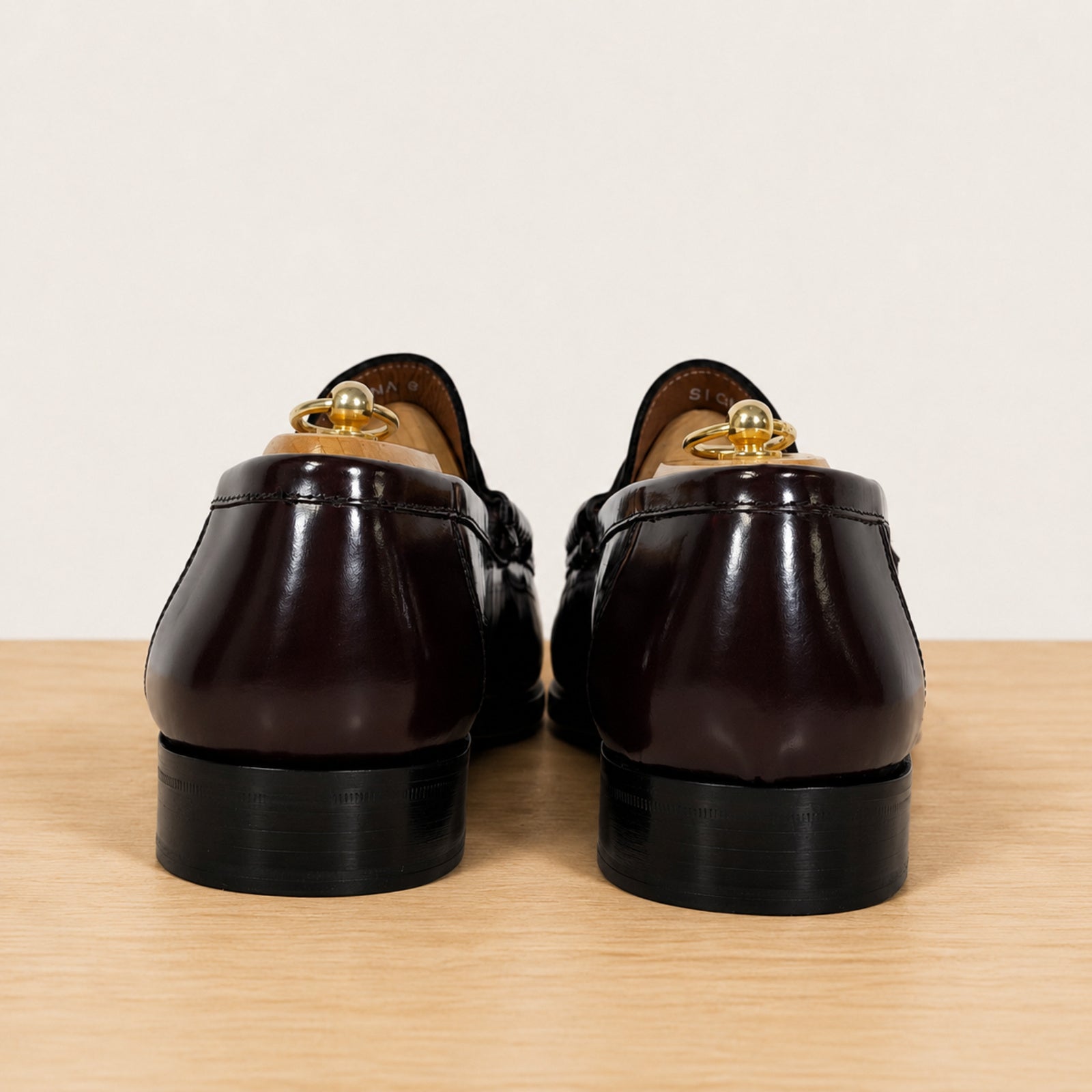 A pair of burgundy patent leather penny loafers, shown from the back on a wooden surface against a light gray background.