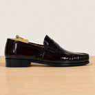 A single burgundy patent leather penny loafer shown in profile on a light wood surface against a warm off-white background.