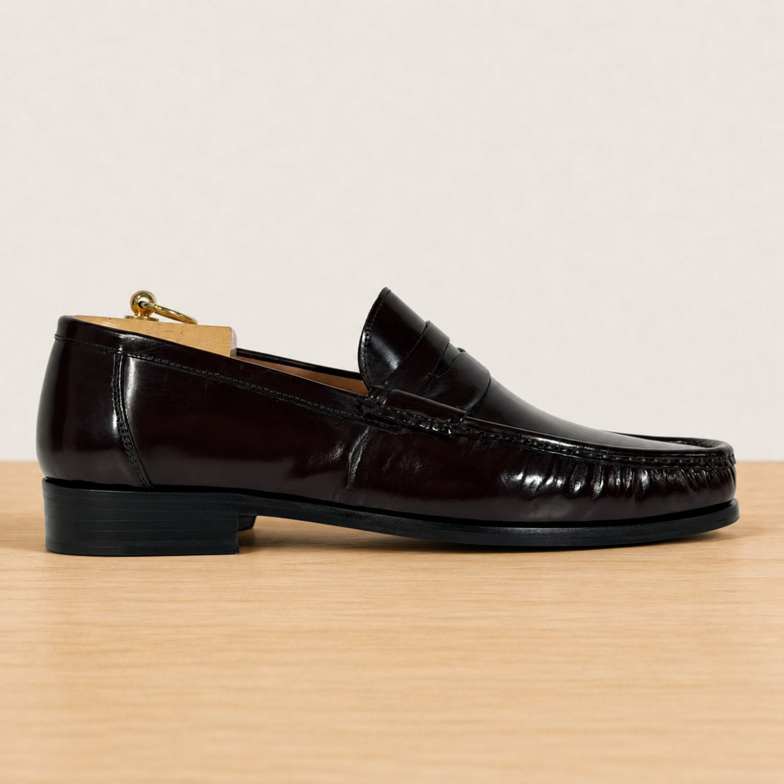 A single burgundy patent leather penny loafer shown in profile on a light wood surface against a warm off-white background.