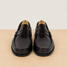 A pair of burgundy glossy leather penny loafers with black soles, displayed on a light wood surface against a neutral off-white background.