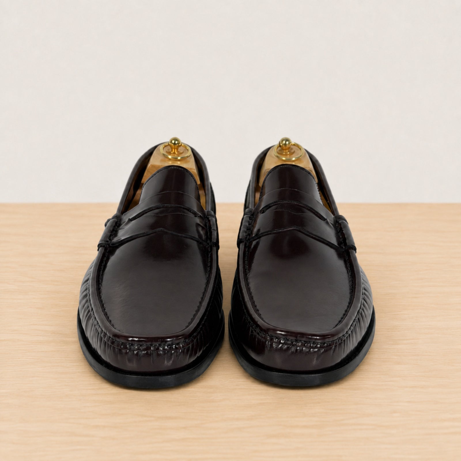 A pair of burgundy glossy leather penny loafers with black soles, displayed on a light wood surface against a neutral off-white background.