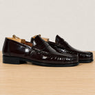 A pair of glossy burgundy penny loafers with a high-shine patent finish, displayed on a wooden surface against a neutral light gray background.