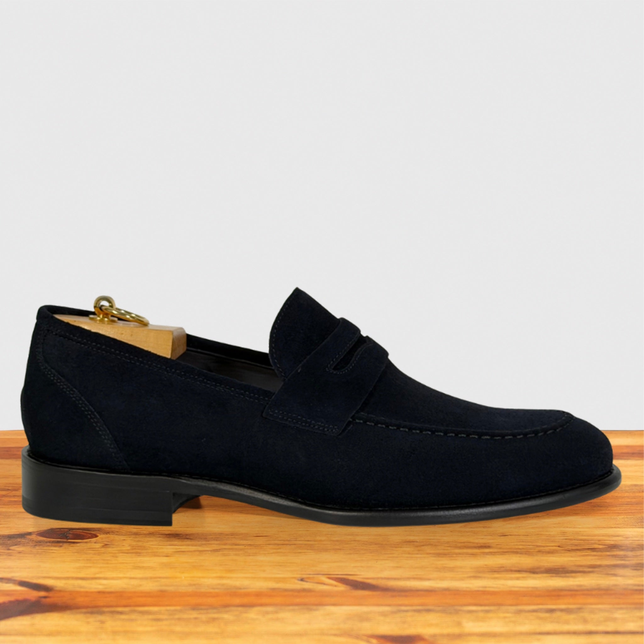 A single blue suede penny loafer shown in profile on a wooden surface against a light gray background.