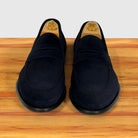 A pair of  blue suede penny loafers displayed on wooden shoe trees on a wooden surface against a white background.