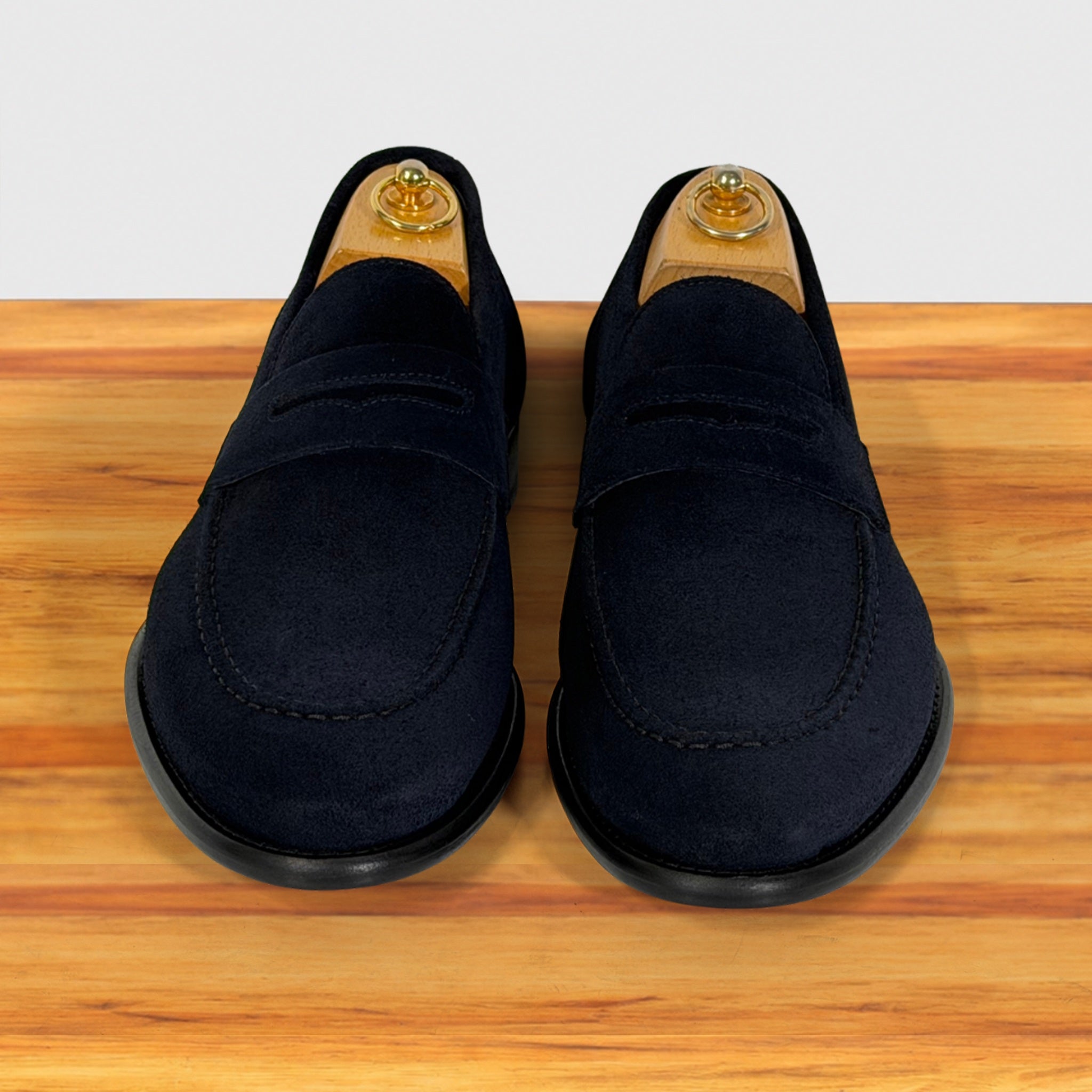 A pair of  blue suede penny loafers displayed on wooden shoe trees on a wooden surface against a white background.