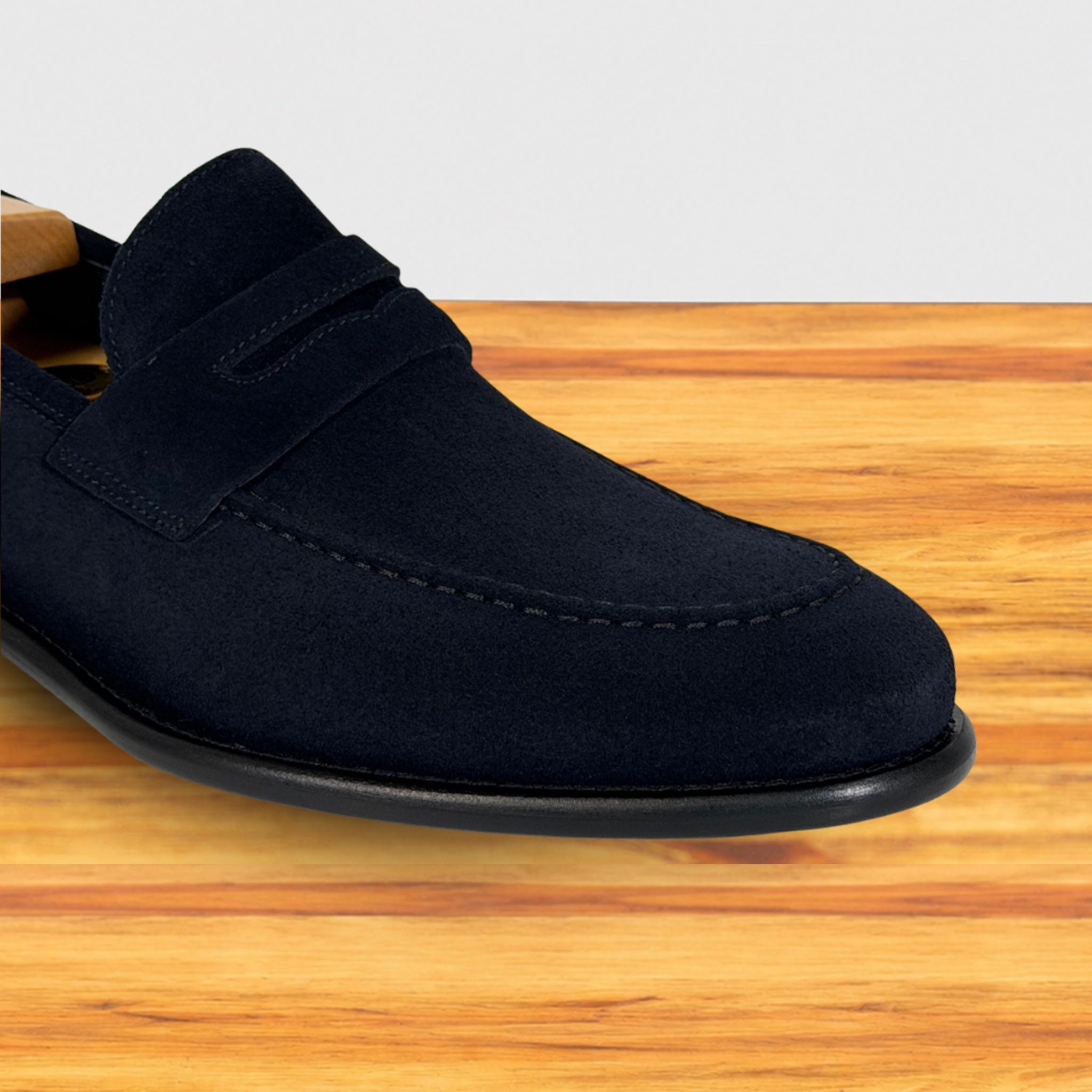 A single  navy blue suede penny loafer shoe resting on a light brown wooden surface against a white background.