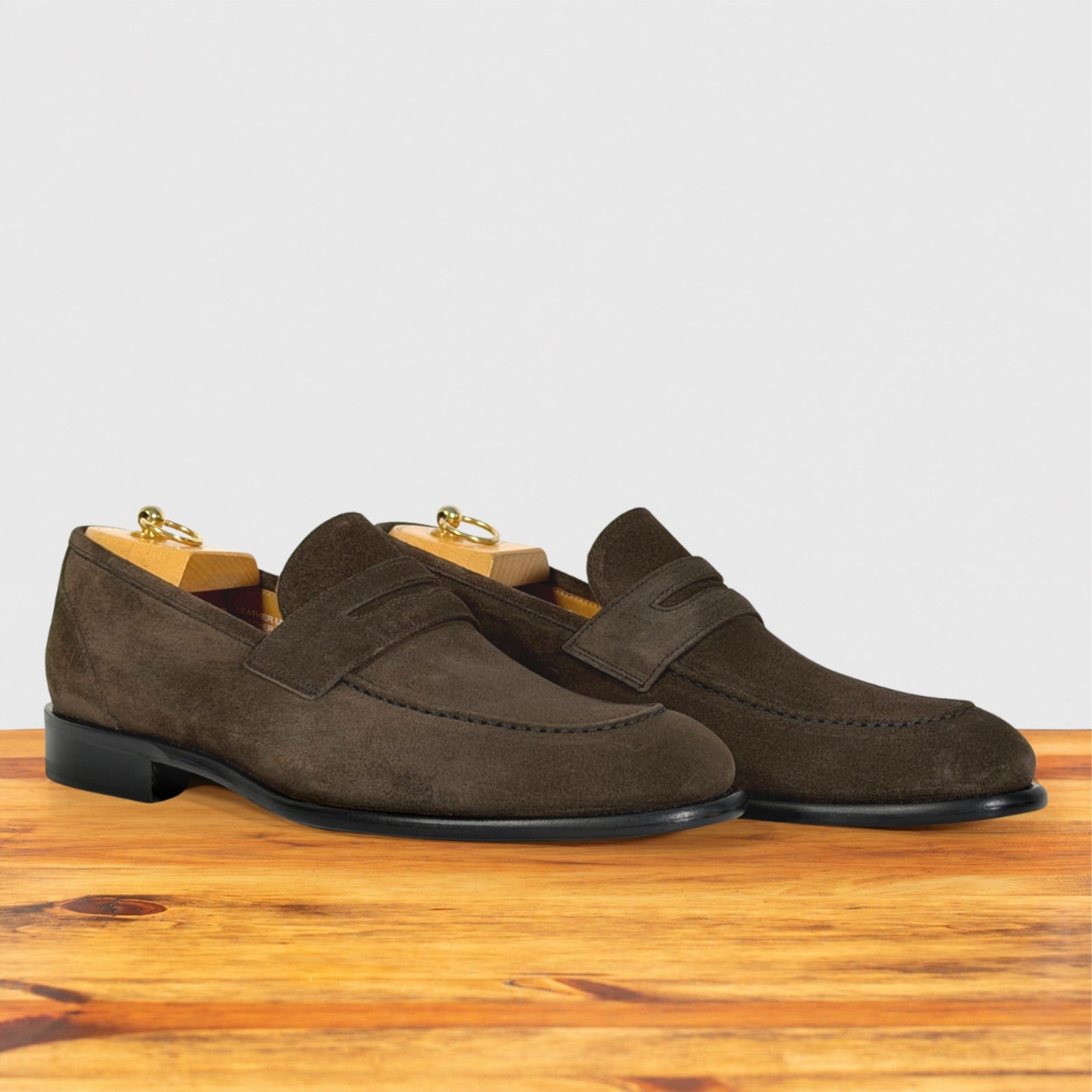 A pair of dusty brown suede penny loafers with black soles, displayed on a wooden surface against a light gray background.