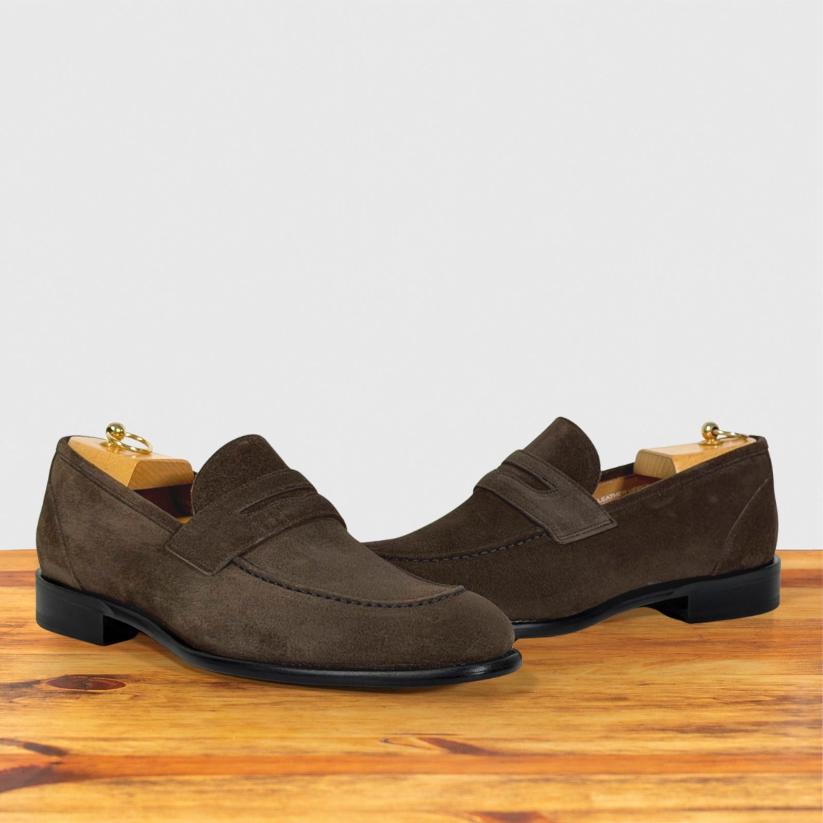 A pair of dusty brown suede penny loafers with black soles, displayed on a wooden surface against a light gray background.