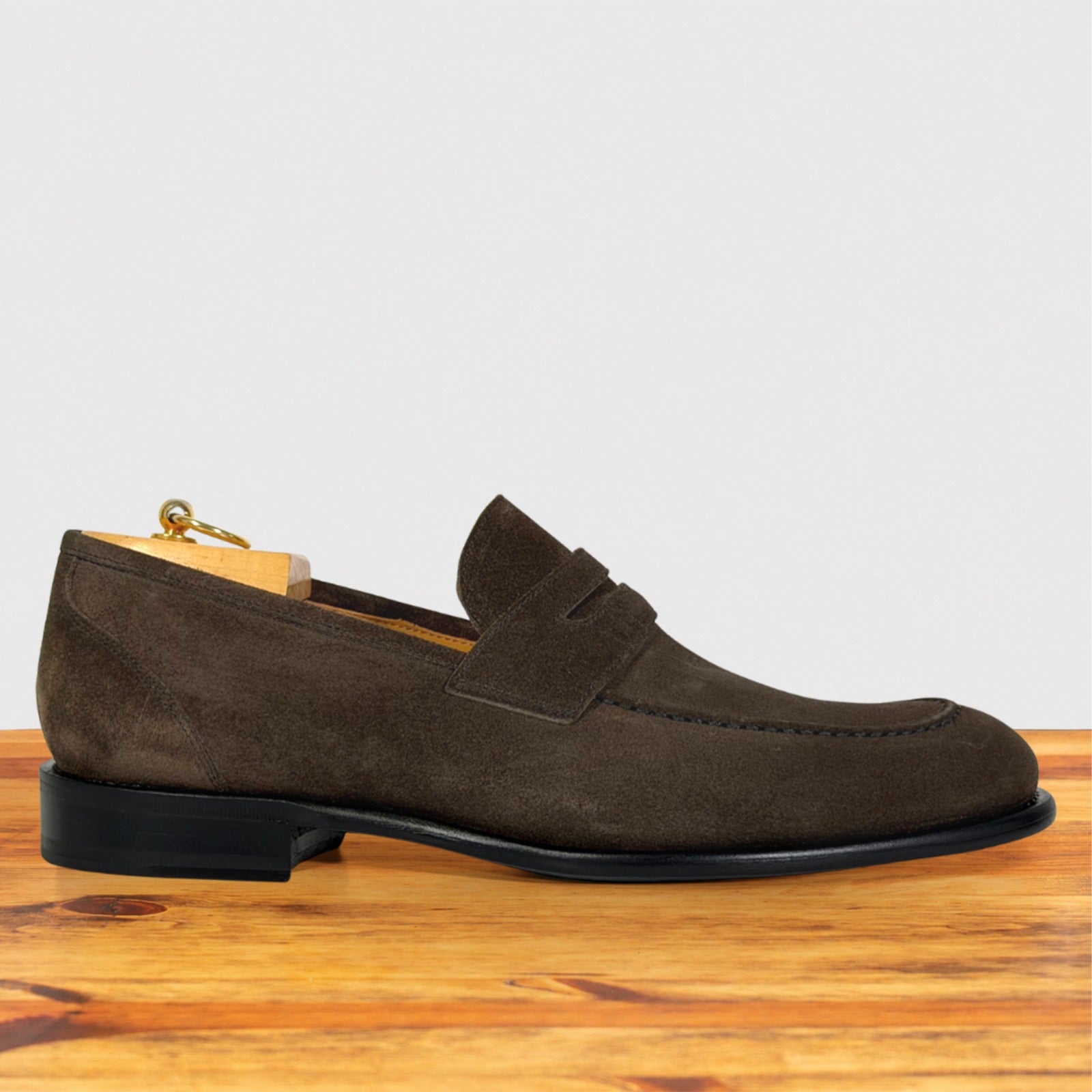 A dusty brown suede penny loafer shoe displayed on a wooden surface against a light gray background.