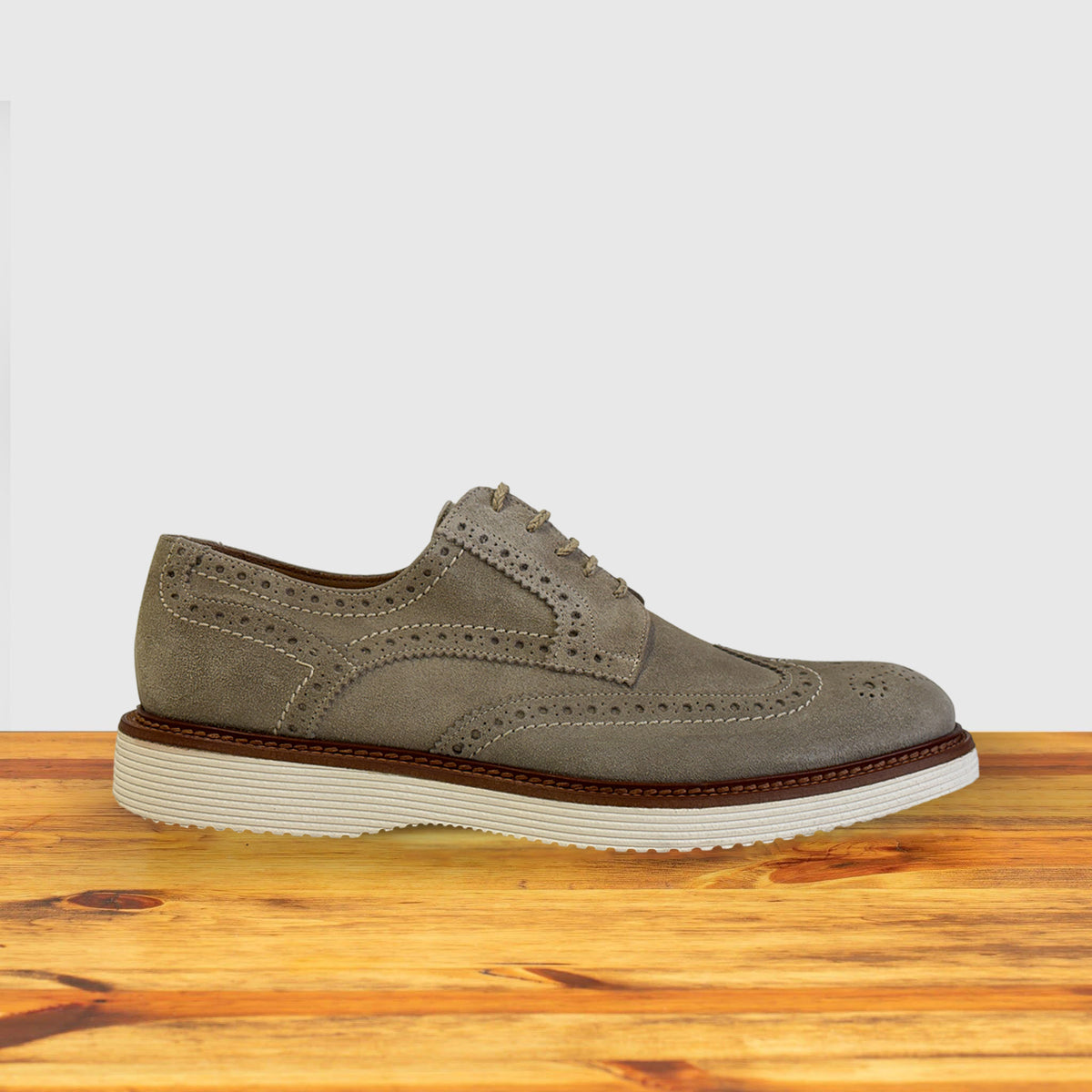 Cashmere suede shoe, side profile view showing the full brogue perforations, wingtip detailing, white ridged midsole on a wooden surface.
