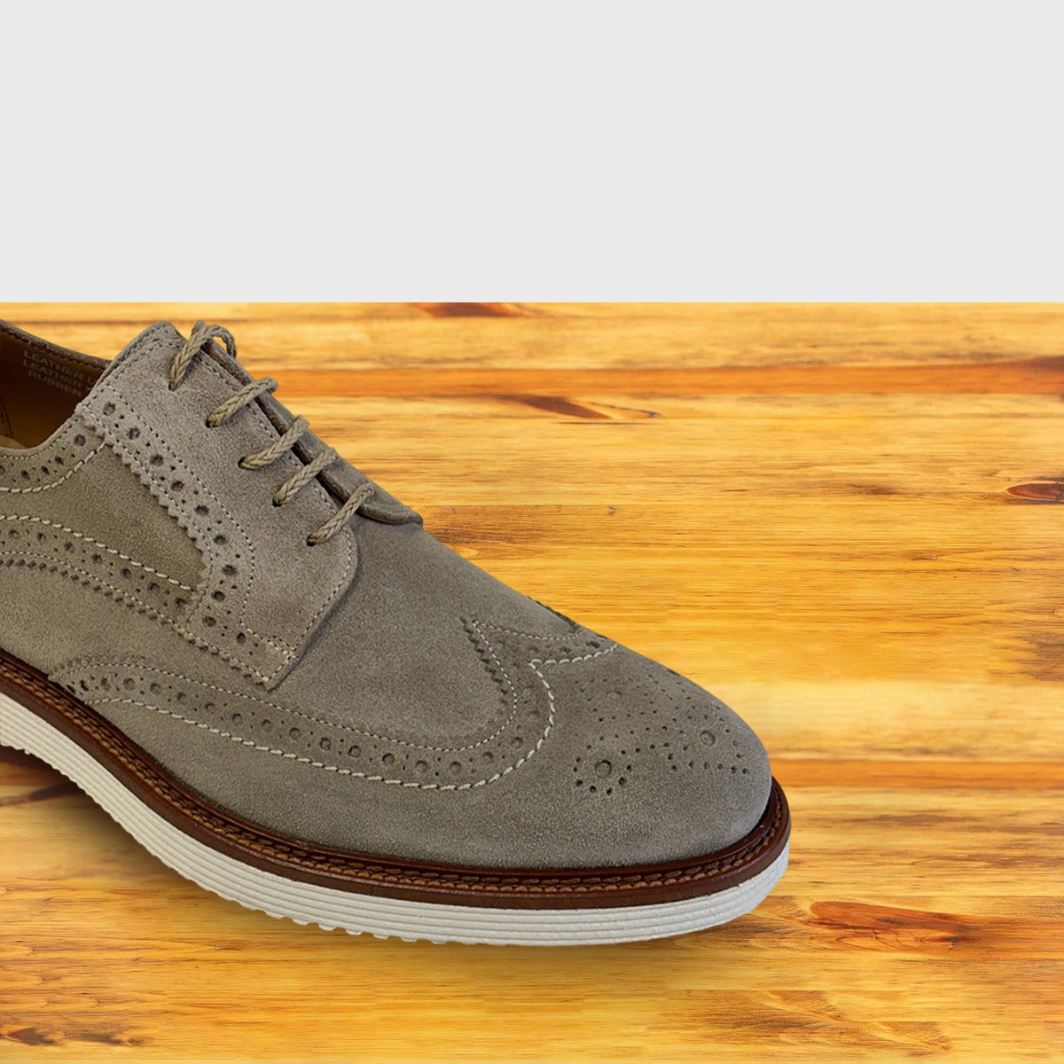 A single men's wingtip oxford, Cashmere color shoe in taupe suede, shown from a side angle on a light-colored wooden surface with a gray background.