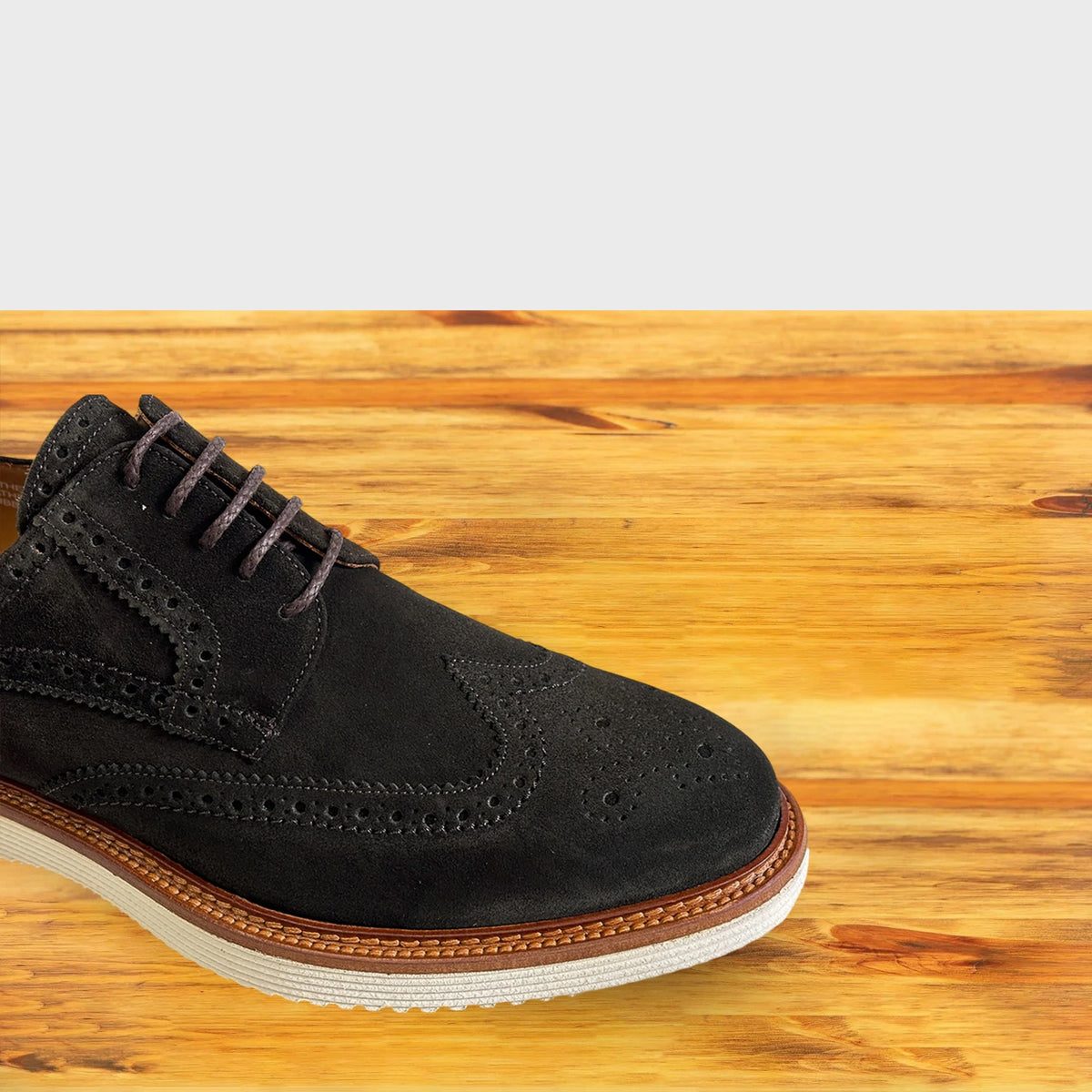 A Olive Gress suede wingtip shoe with laces and a brown welt, resting on a light-colored wooden surface against a gray background.