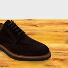 A Chocolate Brown suede wingtip oxford shoe with brogue detailing and laces, positioned on a light-colored wooden surface against a gray background.