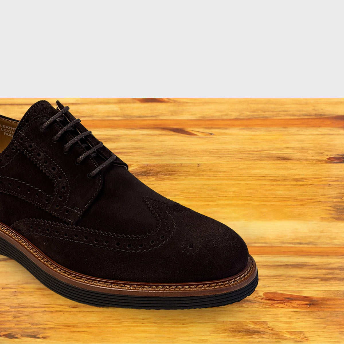 A Chocolate Brown suede wingtip oxford shoe with brogue detailing and laces, positioned on a light-colored wooden surface against a gray background.