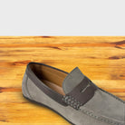 A close-up of a Gray velour suede penny loafer with a leather saddle strap and black sole, shown from a front angle on a wooden table.