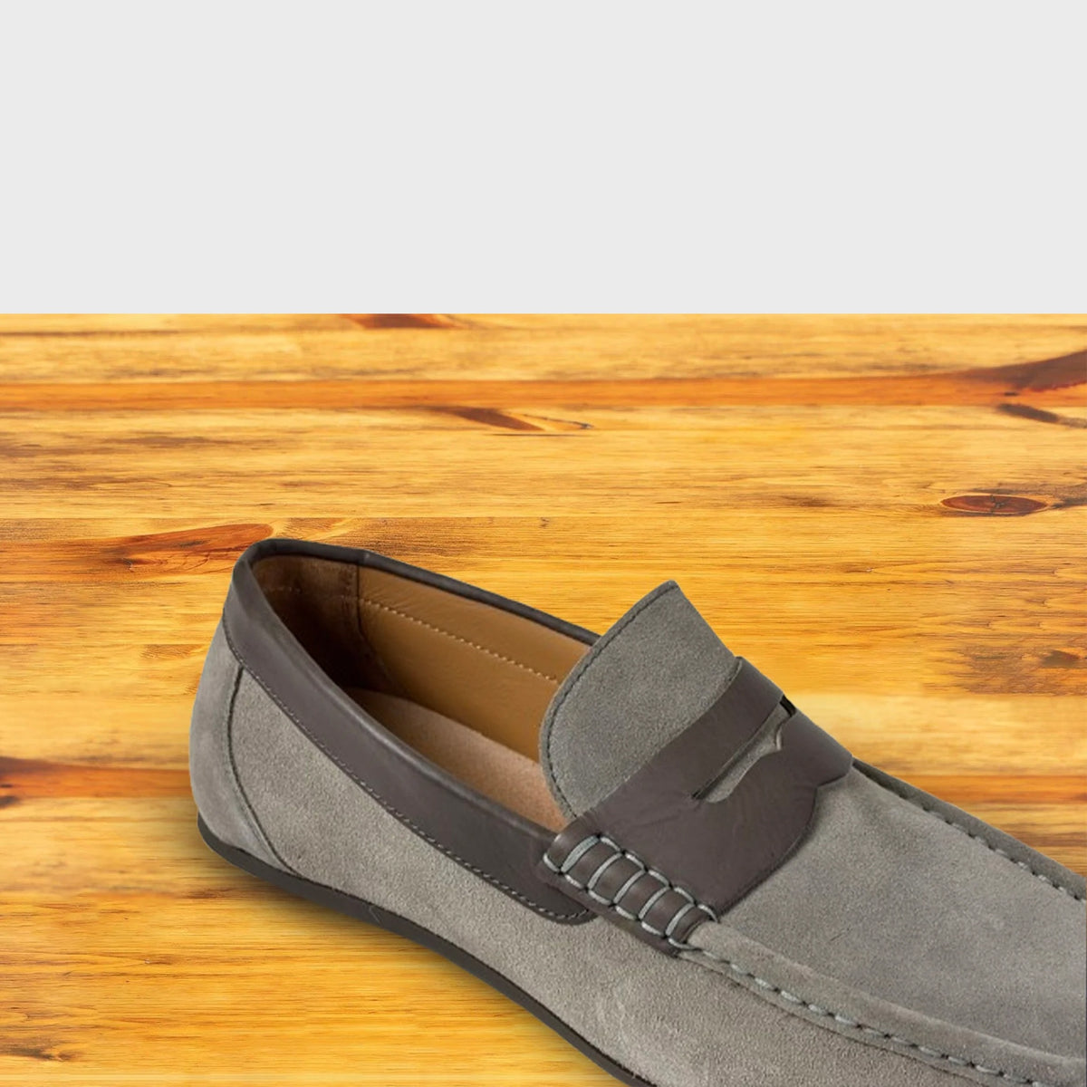 A close-up of a Gray velour suede penny loafer with a leather saddle strap and black sole, shown from a front angle on a wooden table.