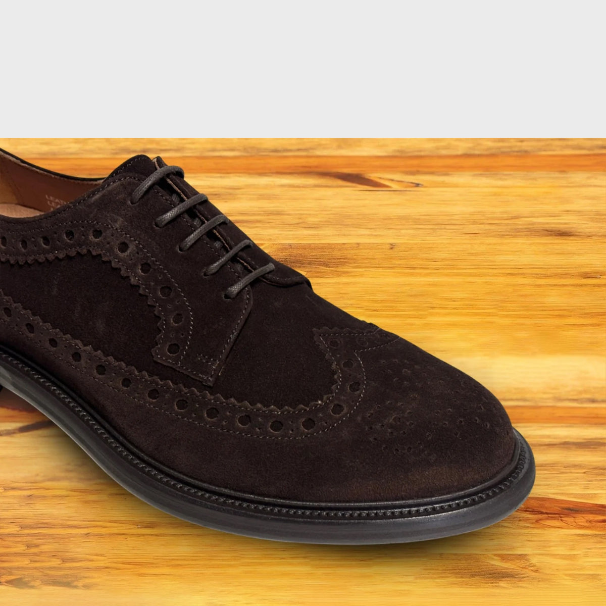 Close view of Chocolate brown suede wingtip shoe with decorative perforations and lace-up closure, displayed on a wooden surface