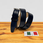 A dark navy blue leather belt with a silver buckle, coiled on a wooden surface next to a small 'Made in Italy' card.