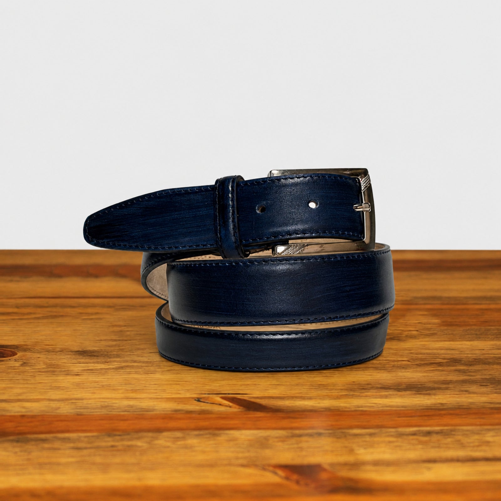 A dark navy blue leather belt with a silver-tone metal buckle, neatly coiled on a rustic wooden surface against a clean white background.