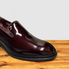 A close-up of a Burgundy patent leather slip-on dress loafer with a black sole, shown from a front angle on a wooden table.