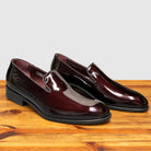 A pair of Burgundy patent leather slip-on dress loafers with black soles, shown from a front three-quarter angle.