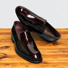 A pair of Burgundy patent leather slip-on dress loafers with black soles, one resting on the other showing the Calzoleria Toscana Made in Italy insole, on a wooden table.
