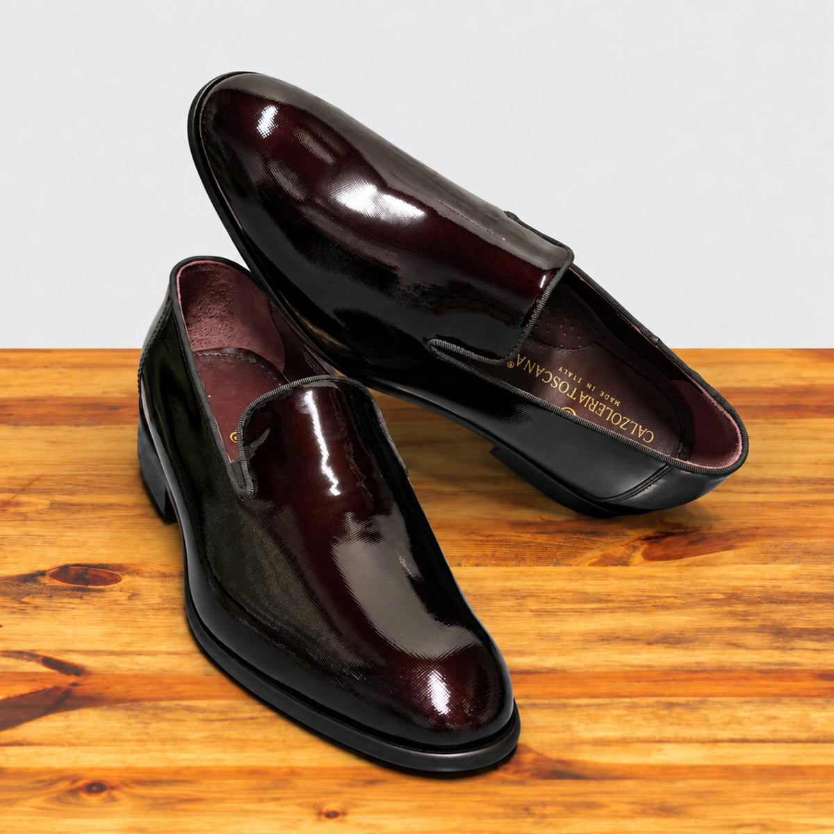A pair of Burgundy patent leather slip-on dress loafers with black soles, one resting on the other showing the Calzoleria Toscana Made in Italy insole, on a wooden table.