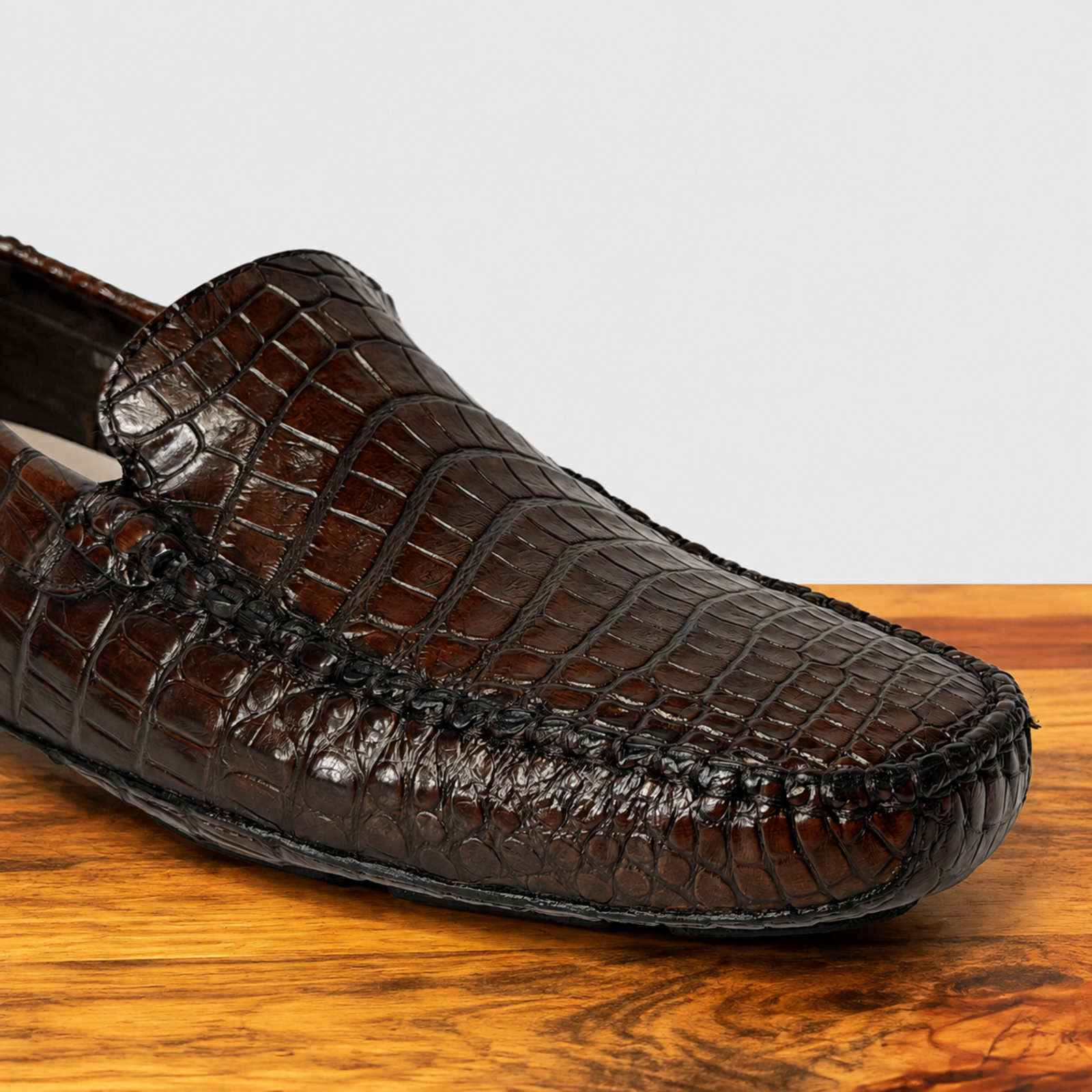 A close-up of a cerris color crocodile-leather loafer on a wooden surface against a light gray background.