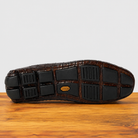 The outsole of a cerris color crocodile- leather shoe, showing black rubber tread blocks and a small gold-tone logo medallion, resting on a wooden surface against a light gray background.