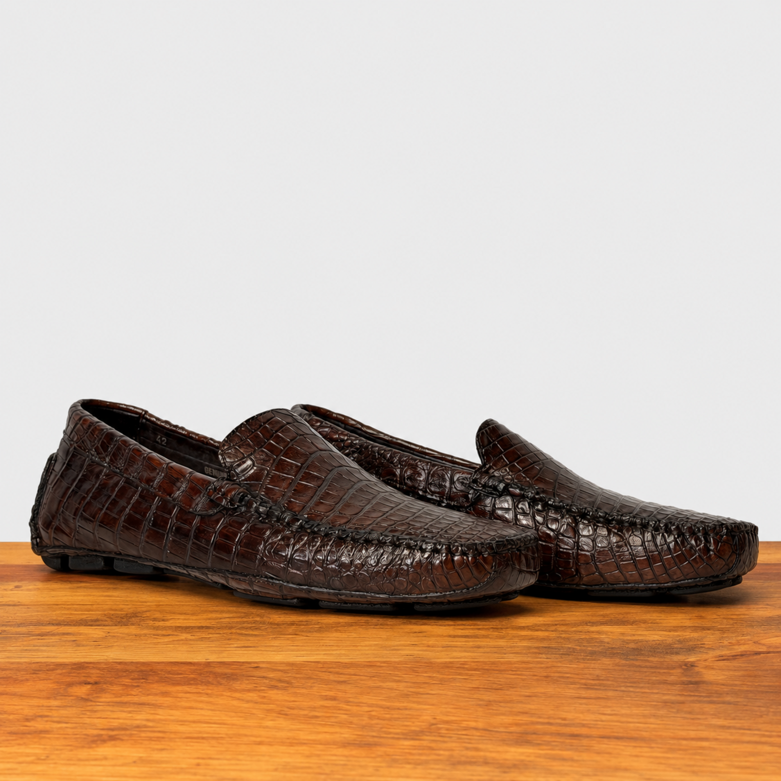 A pair of cerris color crocodile leather driving loafers resting on a polished wooden surface against a light gray background.