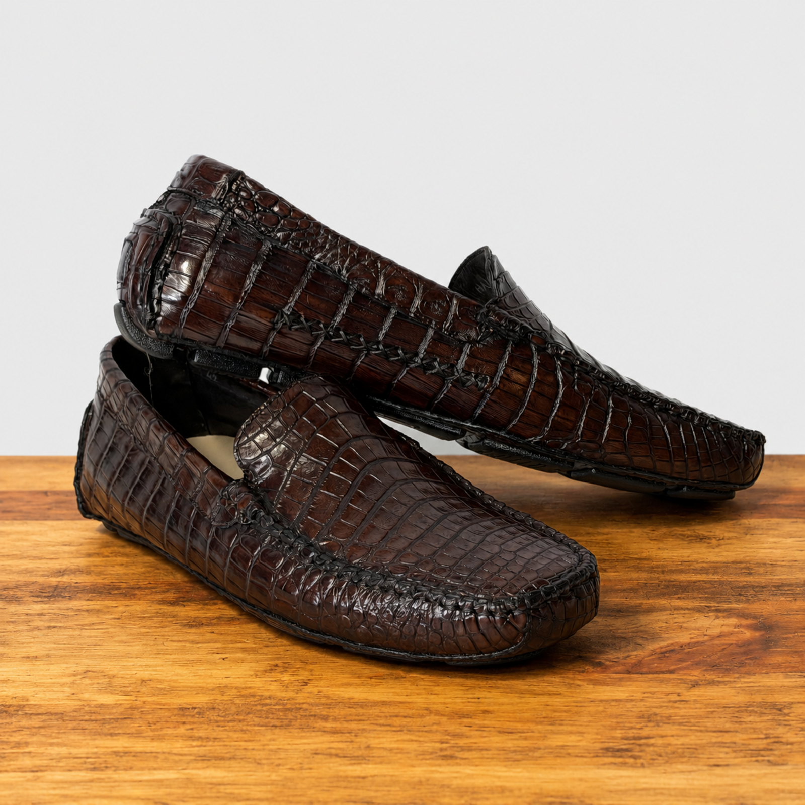 A pair of dark brown crocodile- leather loafers resting on a wooden surface against a light gray background.