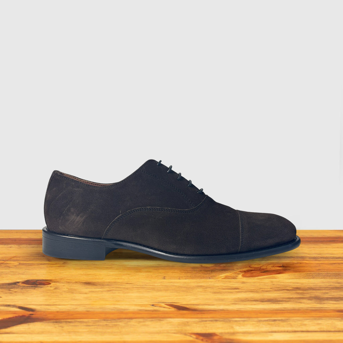 A Dusty Brown color suede Oxford cap-toe shoe with a leather sole, displayed on a wooden surface.