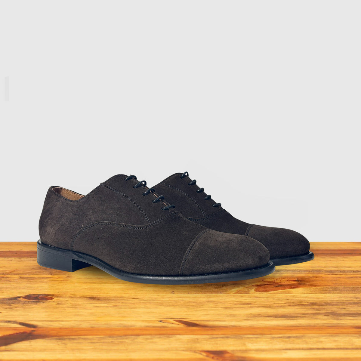 A Pair of Dusty Brown color suede Oxford cap-toe shoe with a leather sole, displayed on a wooden surface.