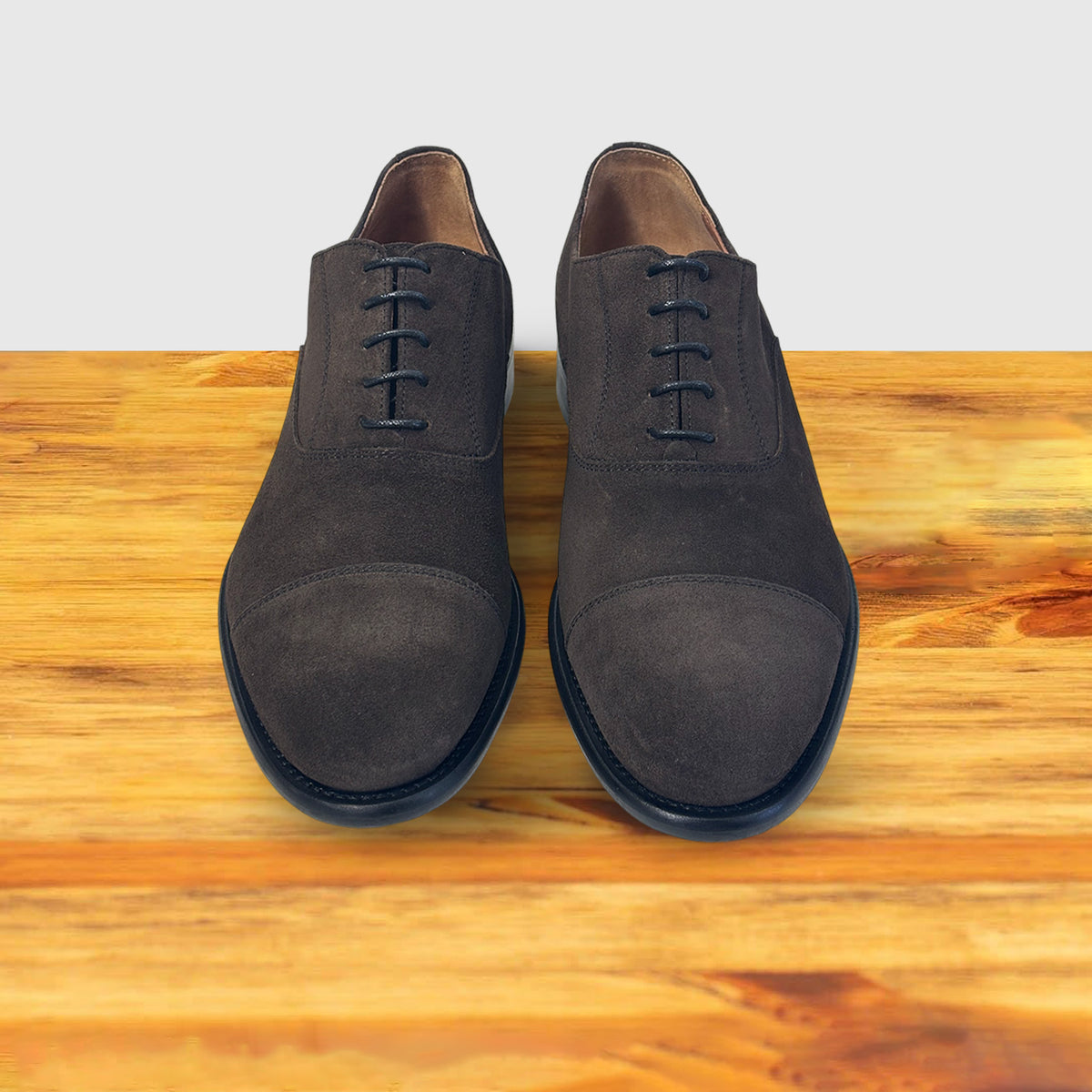A Pair of Dusty Brown Color suede Oxford cap-toe shoe with a leather sole, displayed on a wooden surface.