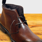 A close-up of a brown leather lace-up ankle boot with dark brown shearling-style lining, shown on a wooden surface.