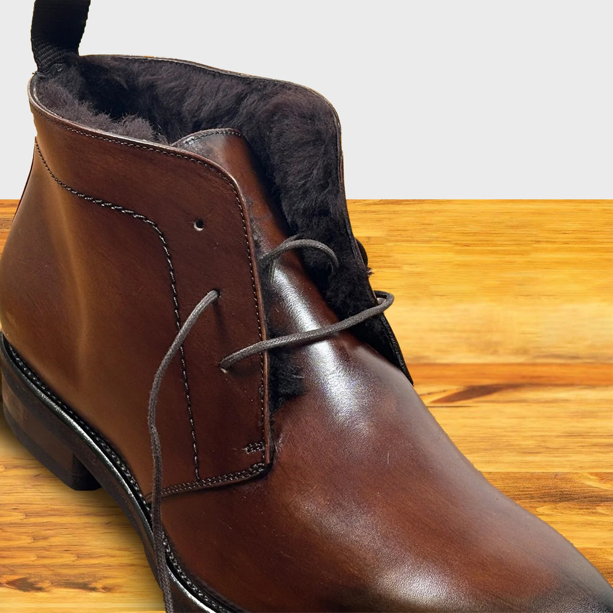 A close-up of a brown leather lace-up ankle boot with dark brown shearling-style lining, shown on a wooden surface.