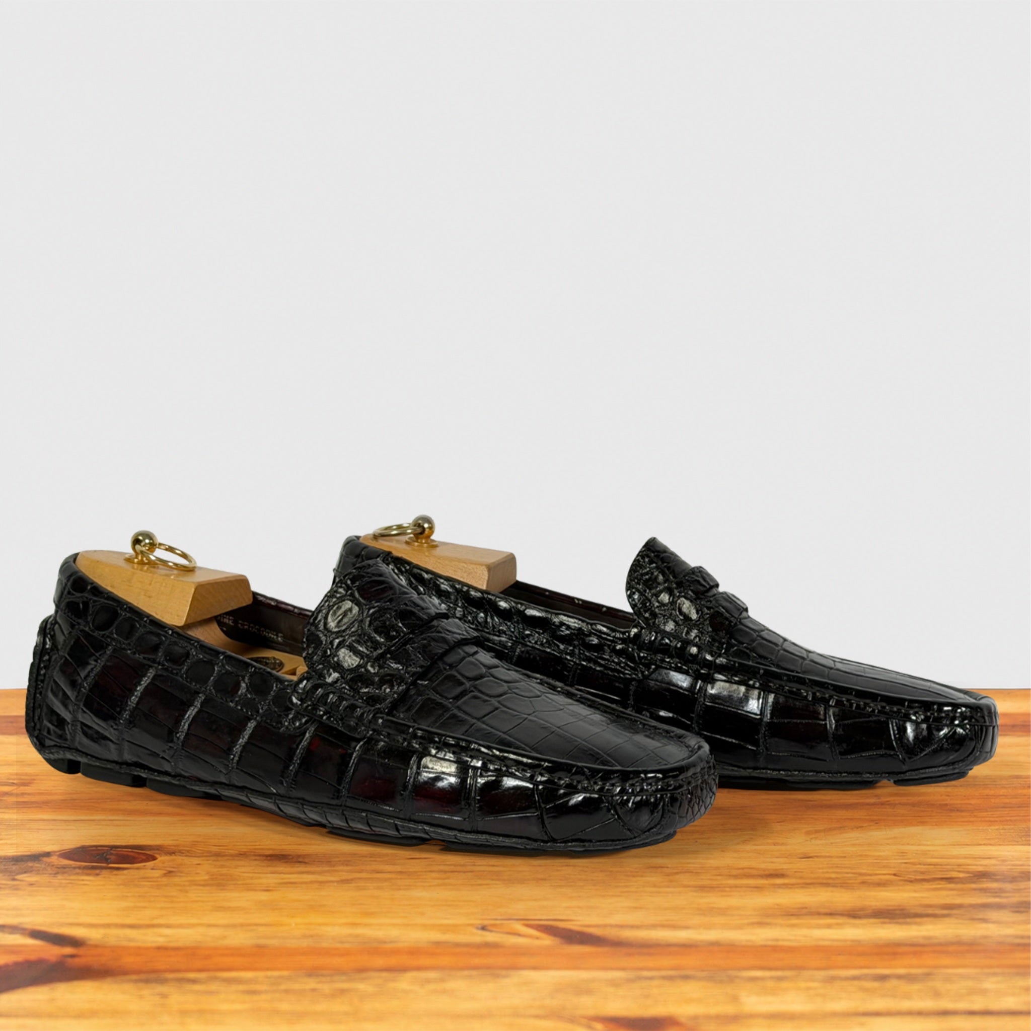 A pair of burgundy crocodile-embossed leather driving loafers with gold-tone metal shoe trees inside, displayed on a wooden surface against a light gray background.