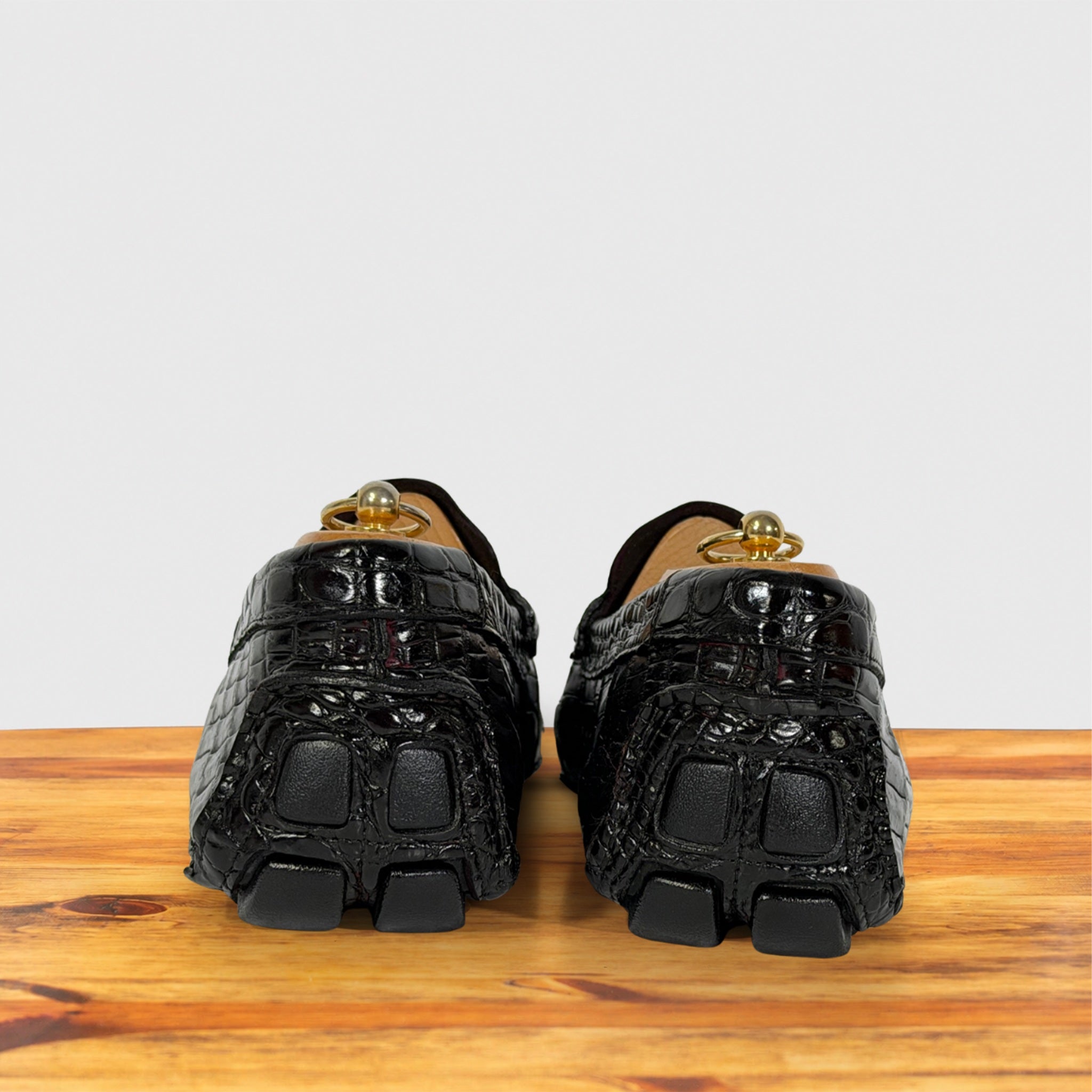 A pair of burgundy crocodile-embossed loafers with chunky block soles , shown from the back on a wooden surface against a light gray background.