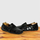 A pair of burgundy crocodile-embossed leather penny loafers, displayed on wooden shoe trees on a wooden surface against a light gray background.
