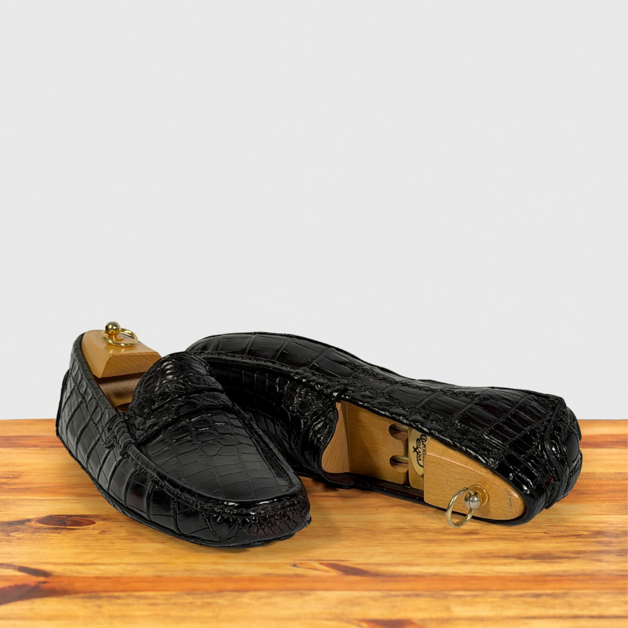 A pair of burgundy crocodile-embossed leather loafers displayed on a wooden surface against a light gray background.